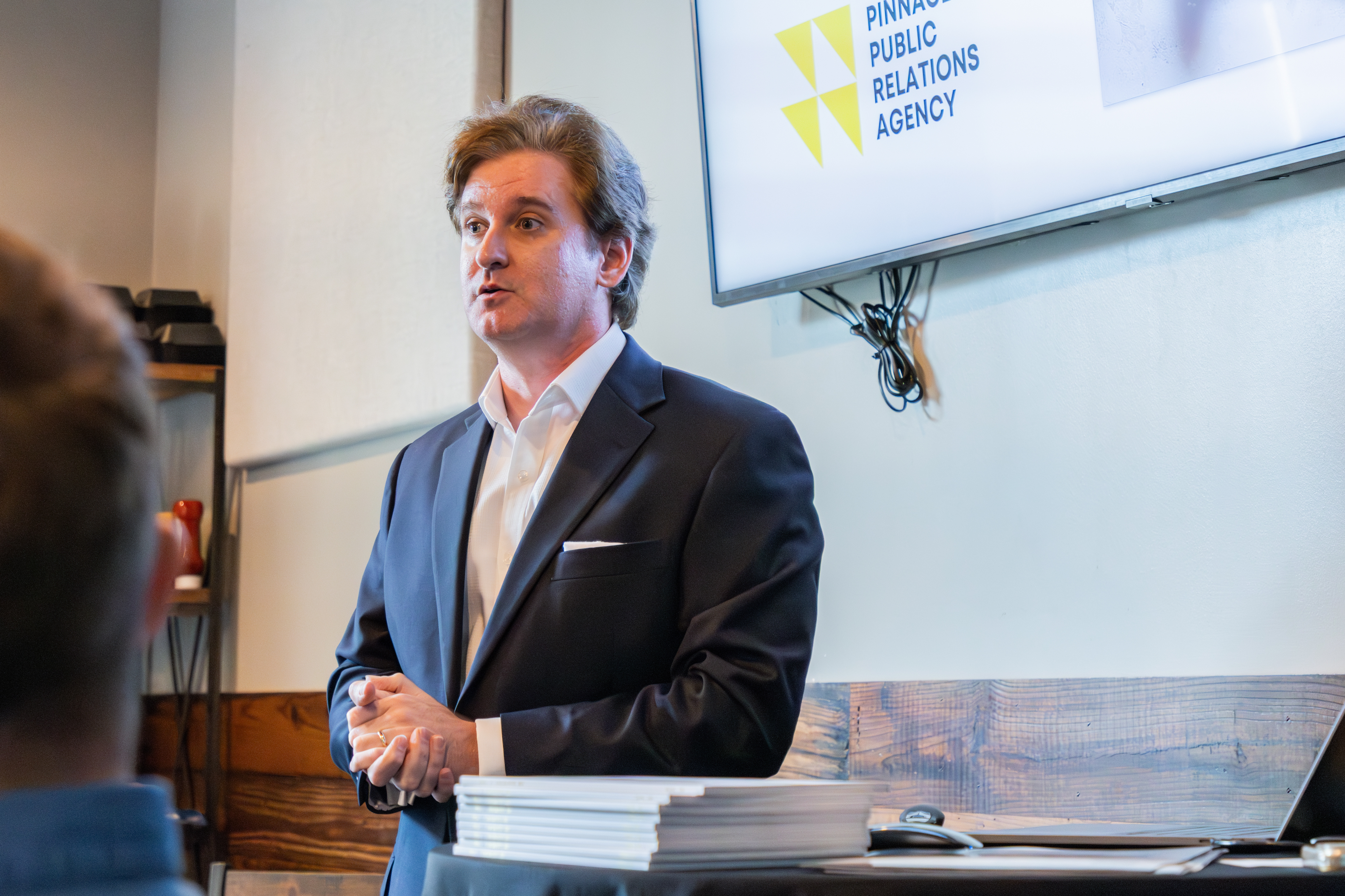 A speaker stands in front of a screen displaying the logo of Pinnacle Public Relations Agency, presenting to an audience in a professional setting.